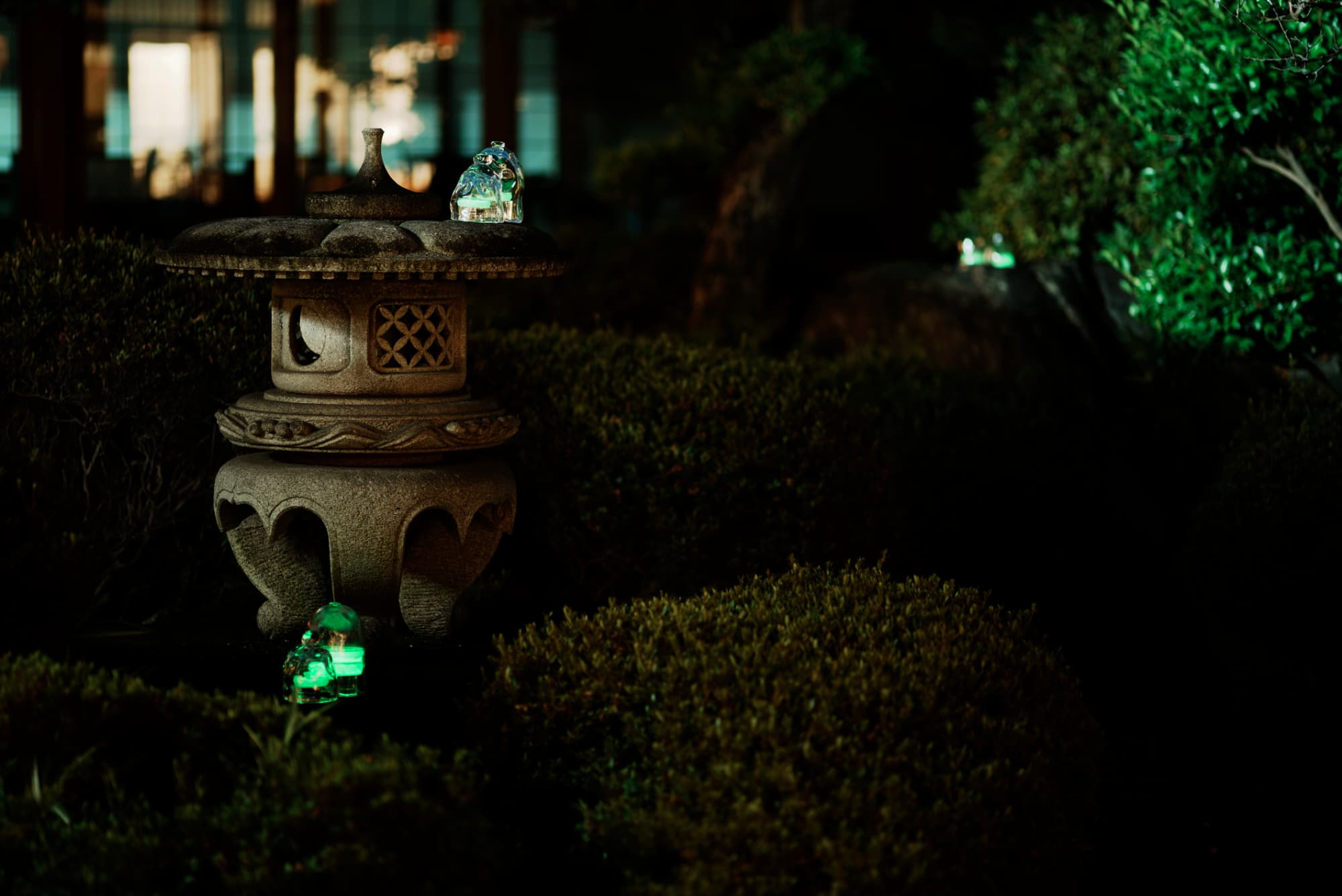 夜の庭の石灯籠のそばで、緑に光るガラスのランプが植栽を照らしている